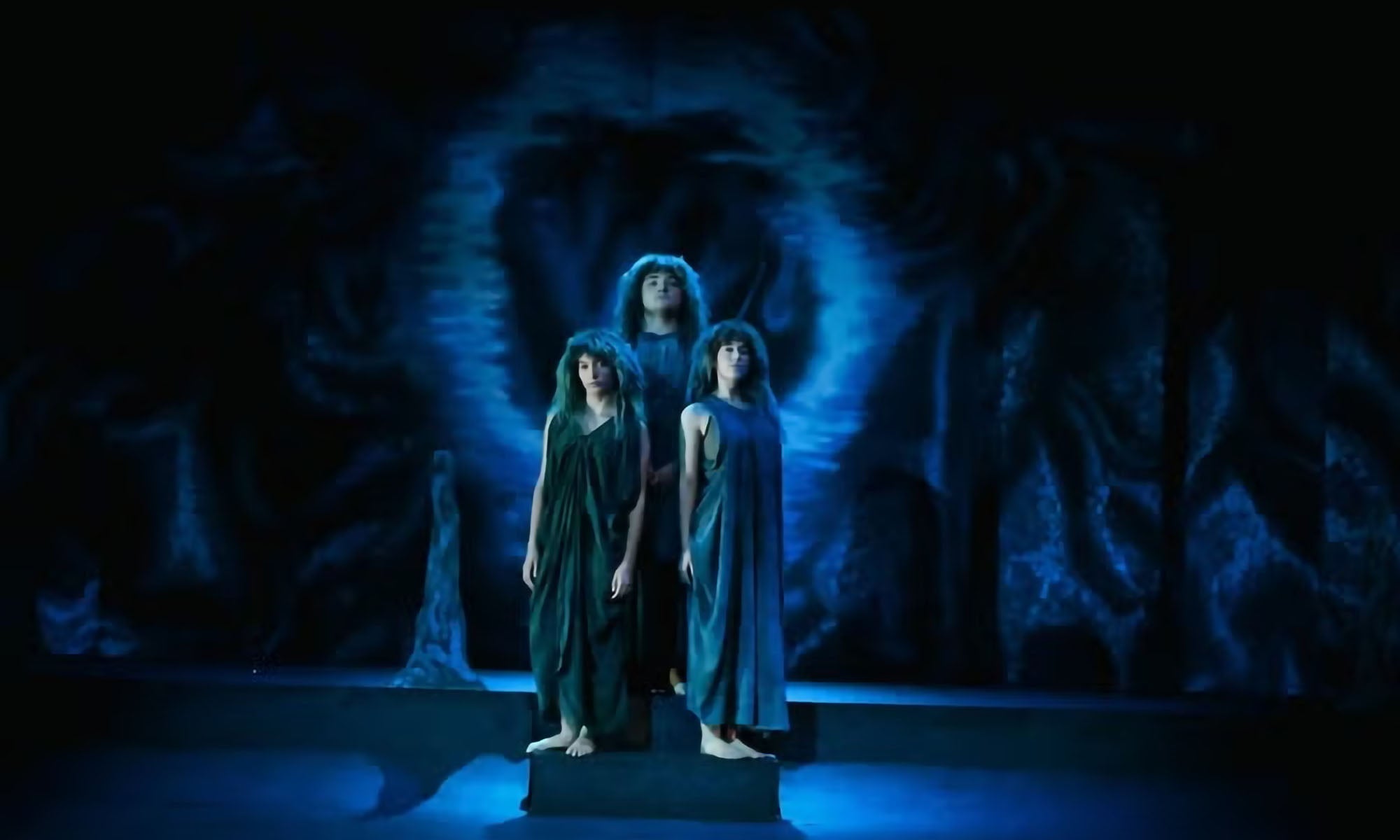 Three actors in gray cloaks clustered together on a stage awash in blue light during a performance of Eurydice.