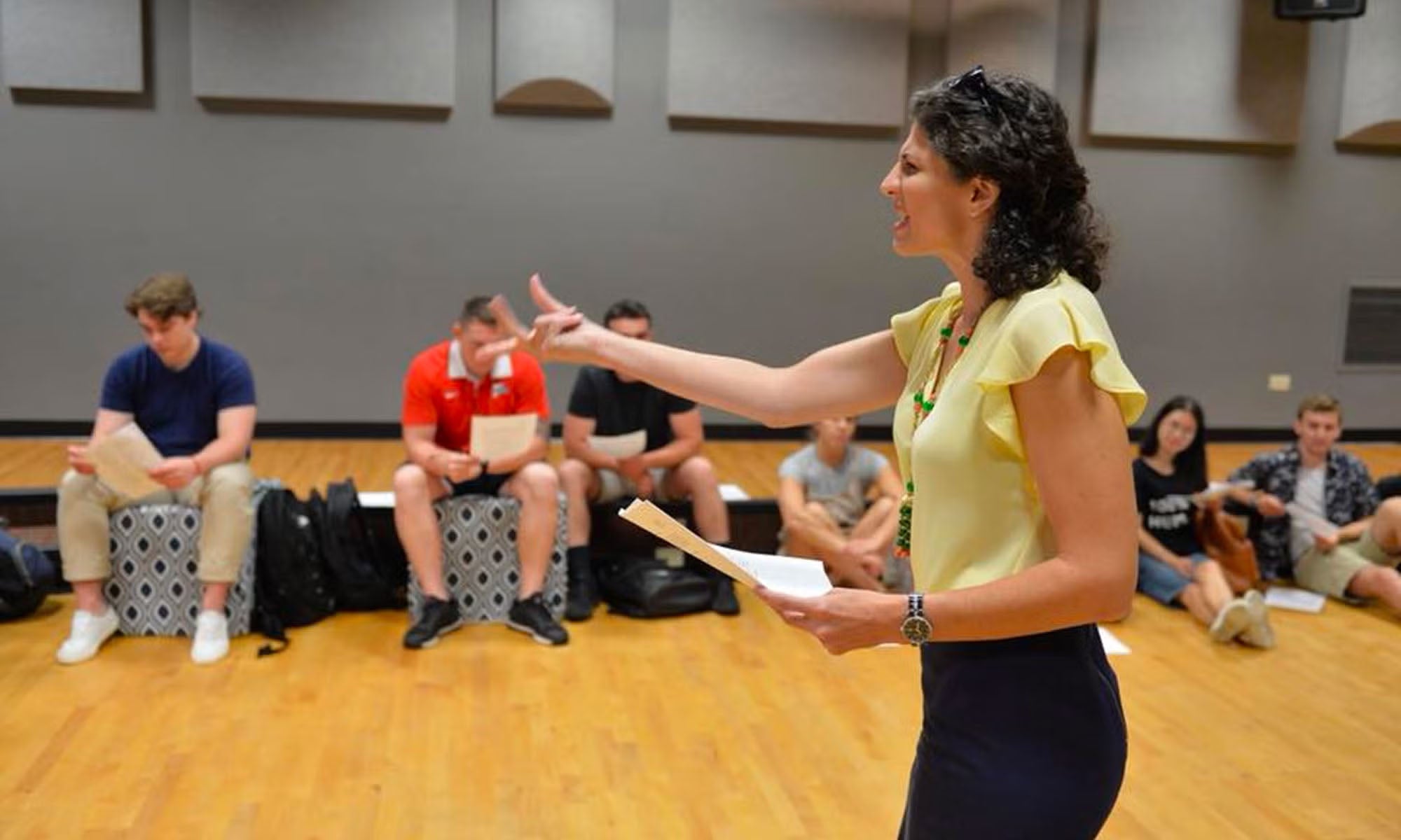 A woman leads a class of actors in a theatrical rehearsal space.