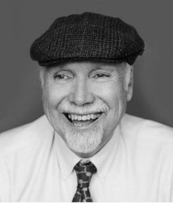 Black and white photo of Gary with white hair and a goatee, smiling. He's wearing a white button down shirt and patterned tie, and a tweed cap.