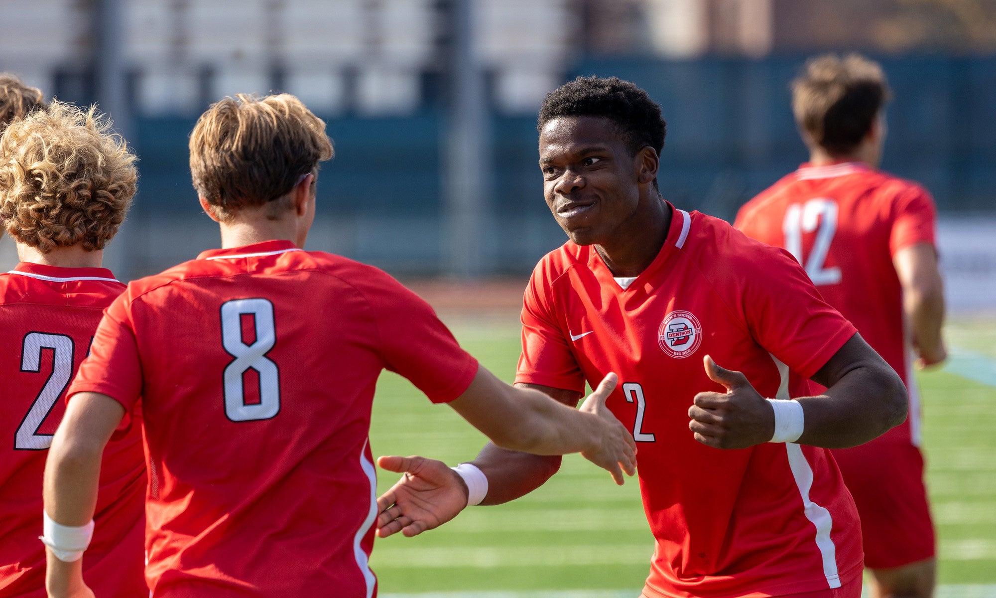 Two soccer players in red jerseys give each other a handshake on the field while teammates gather nearby. The player facing the camera wears No. 2.