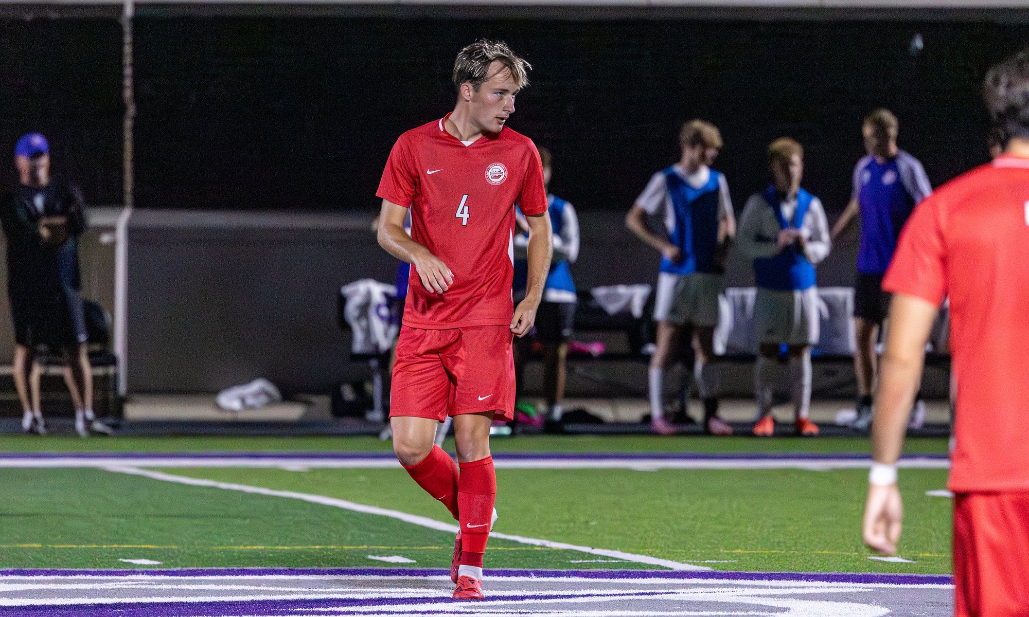 A college soccer player in a red uniform on the field during a night game.