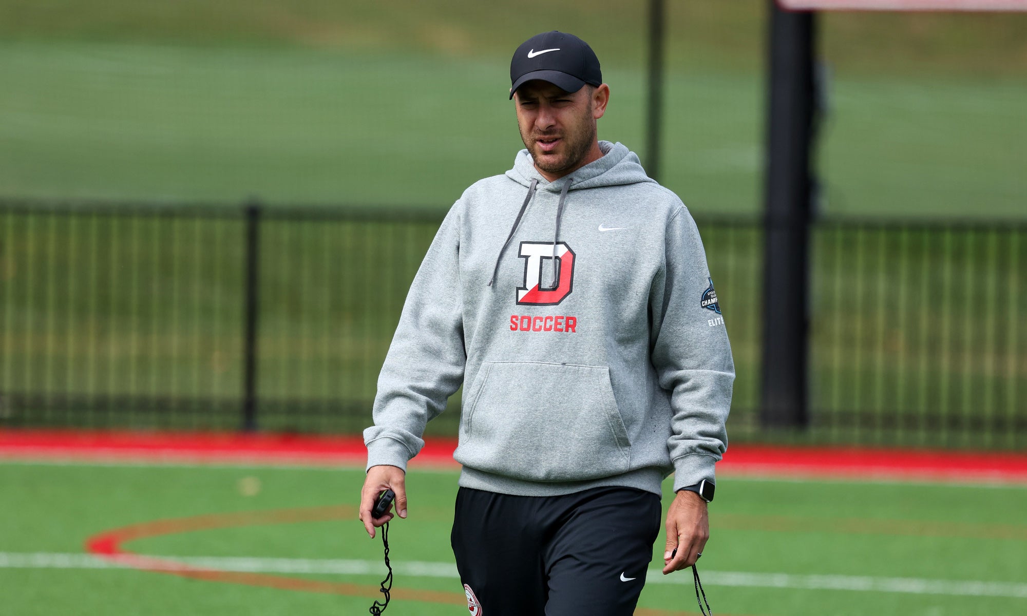 A man in a gray Denison Soccer hoodie and dark ball cap on a soccer field.