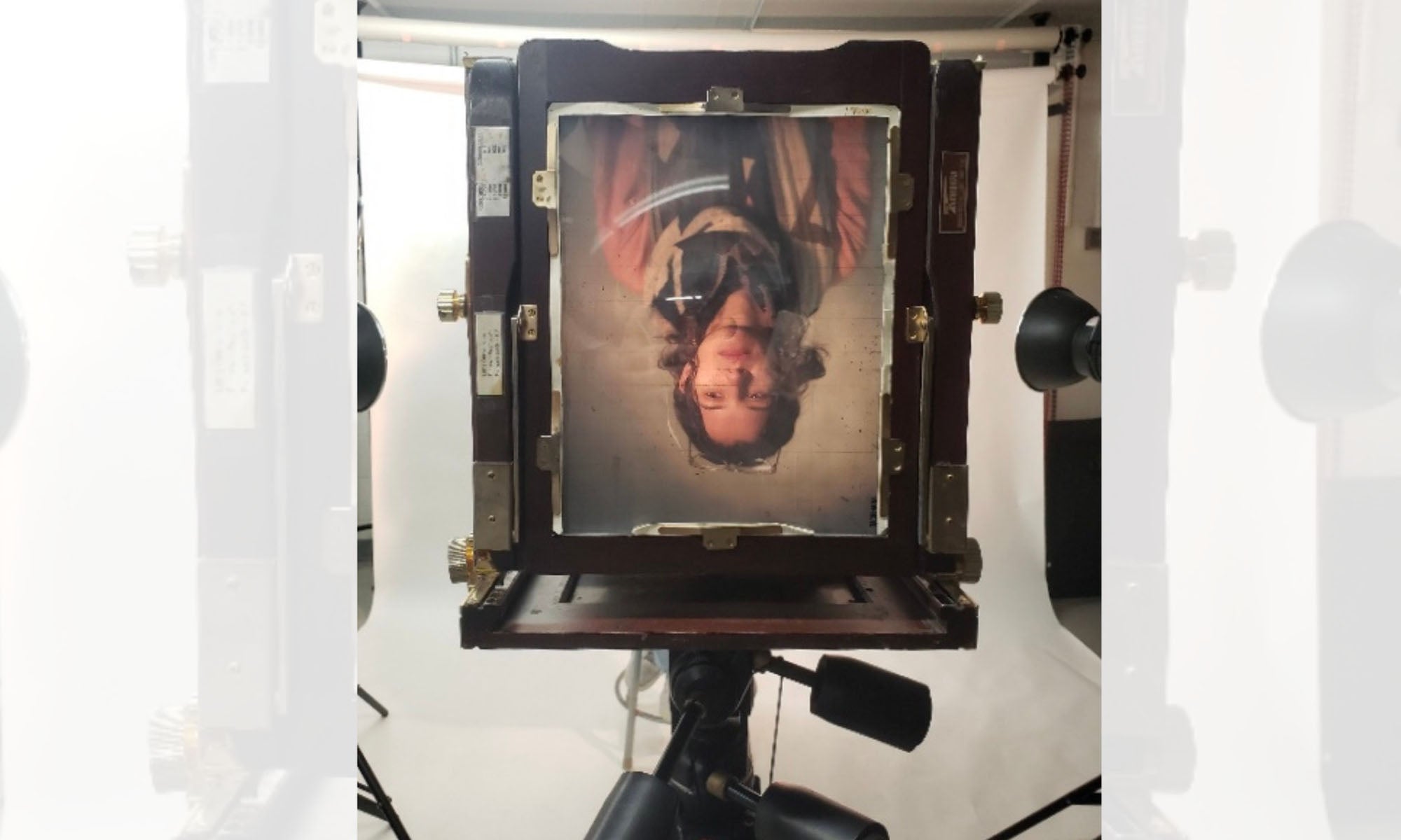 A large-format camera displays an upside-down portrait of a person with long hair and a salmon-pink long-sleeve shirt and brown and beige scarf on its ground glass focusing screen, with studio lights in the background.