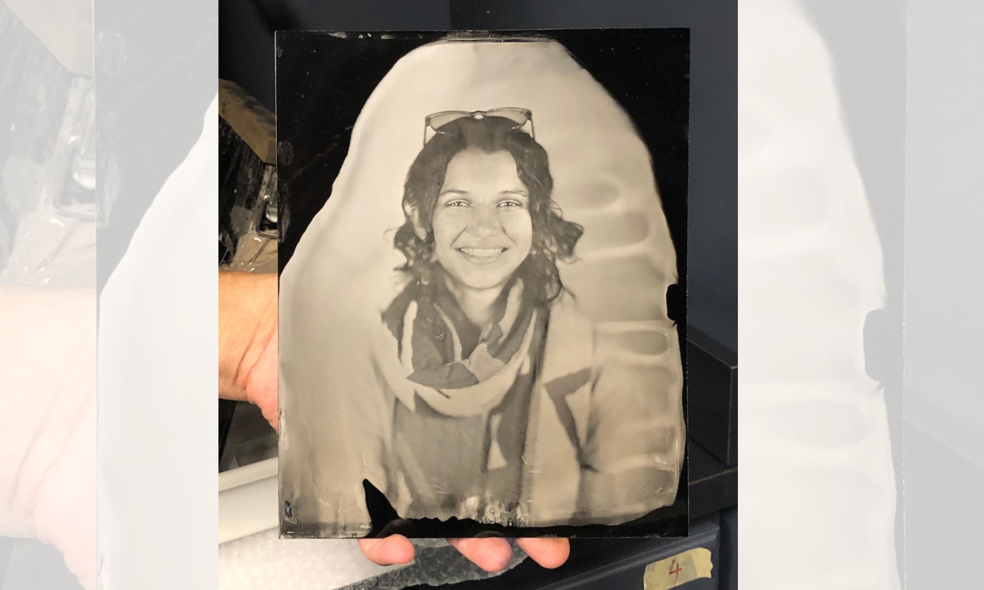 Image of a light sepia-toned tintype of a smiling woman with long dark hair and glasses perched on the top of her head. She wears a long-sleeve shirt and a scarf.