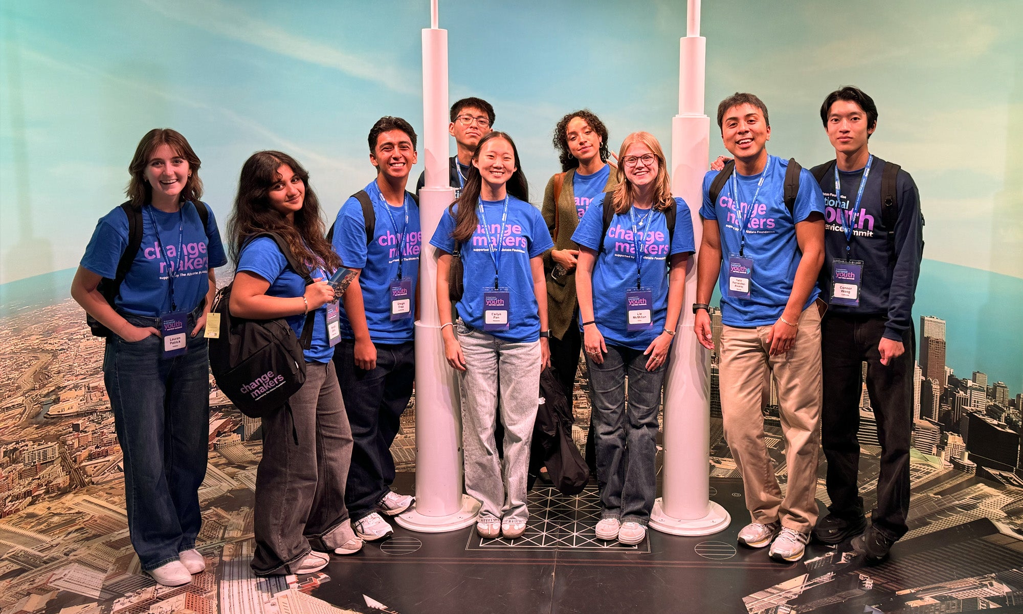 A group of young people wearing matching blue "changemaker" t-shirts and name badges pose together in front of a cityscape backdrop with tall white pillars.