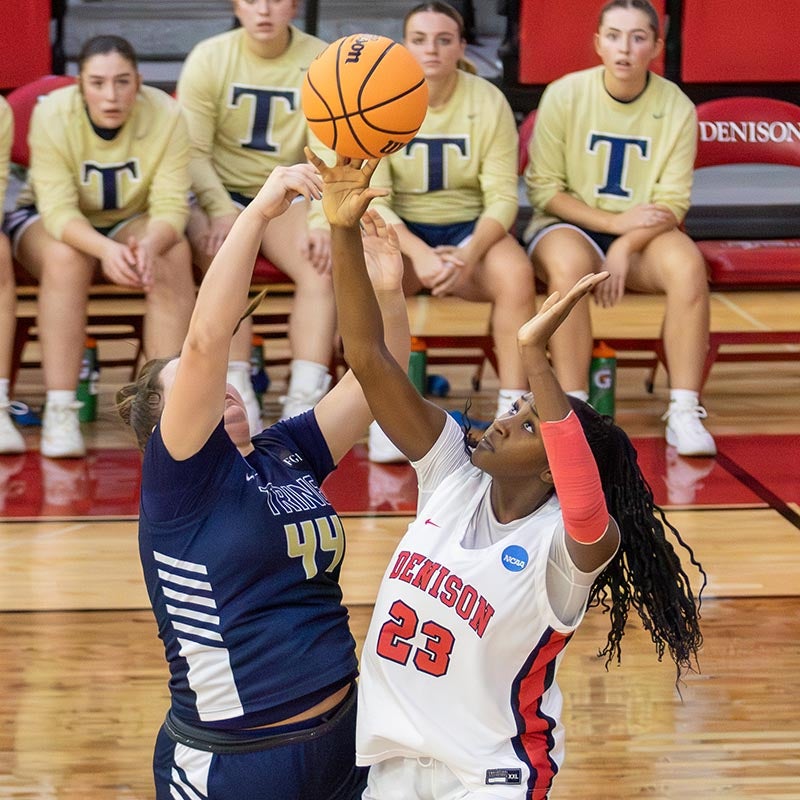 A basketball player in a white Denison jersey, number 23, jumps to shoot as a player in a navy jersey, number 44, tries to block. Four people sit on the bench in the background wearing tan shirts with a T on them.