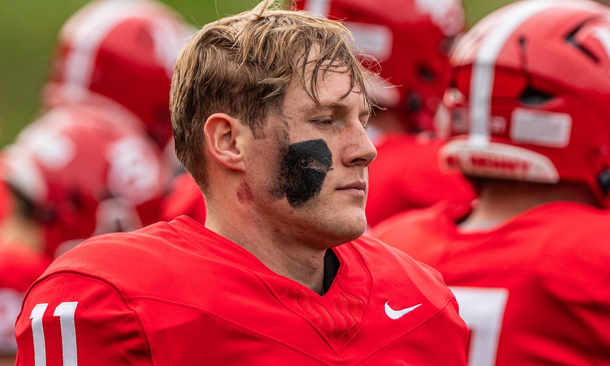 A football player in a red uniform with black eye paint stands on the field with teammates in the background.
