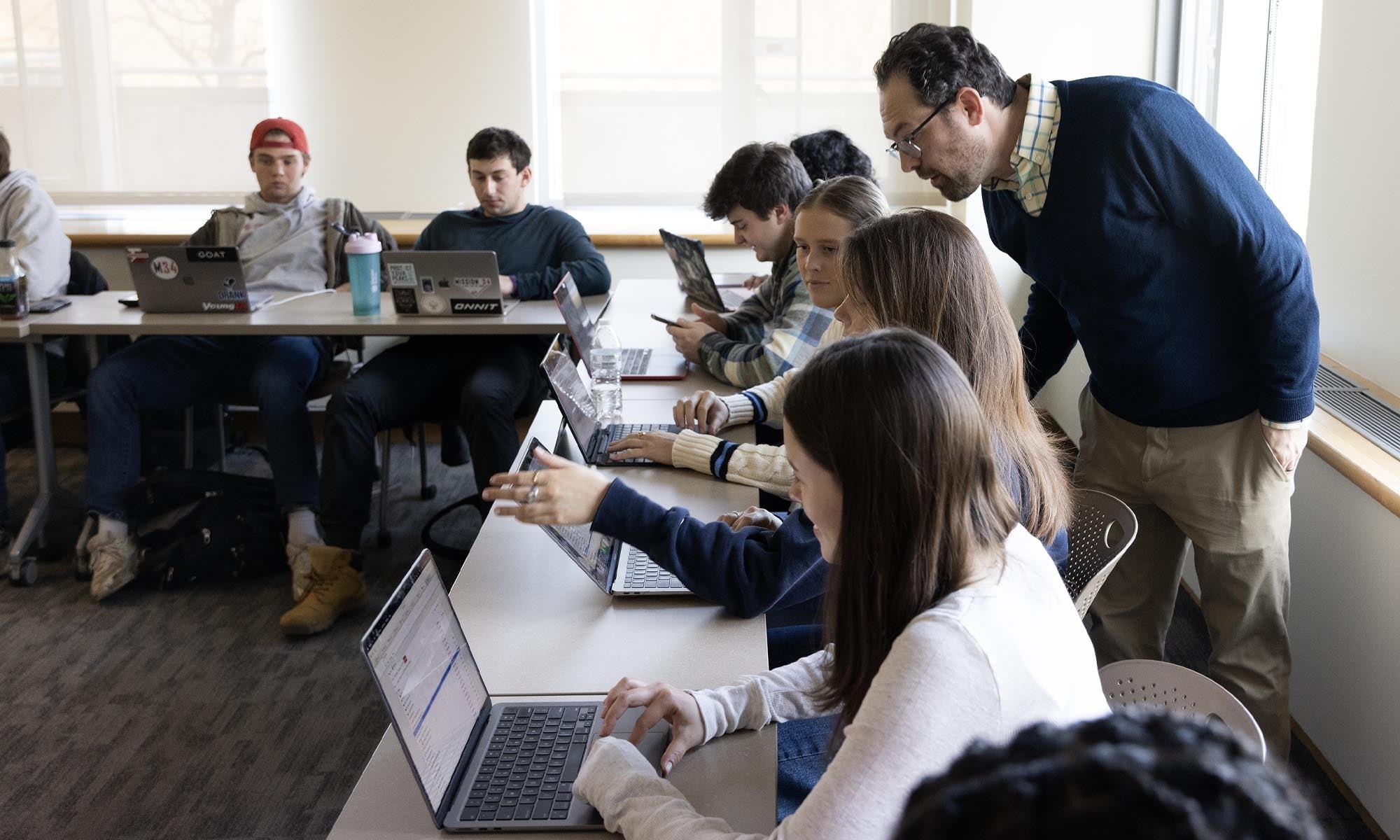 A classroom of student sitting around tables in a conference U-shape configuration working at their laptops; a professor stands nearby answering questions.