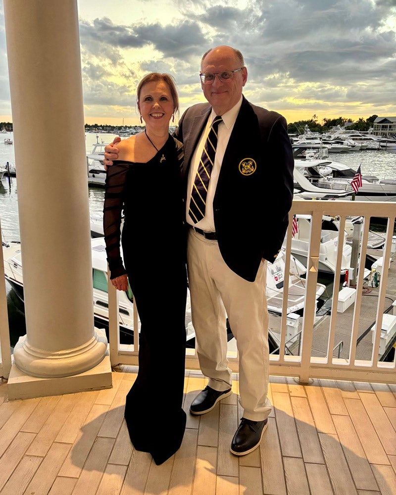 Two people pose together on a balcony overlooking boats at a marina during sunset; the woman wears a black dress and the man wears a blazer, tie, and light pants.