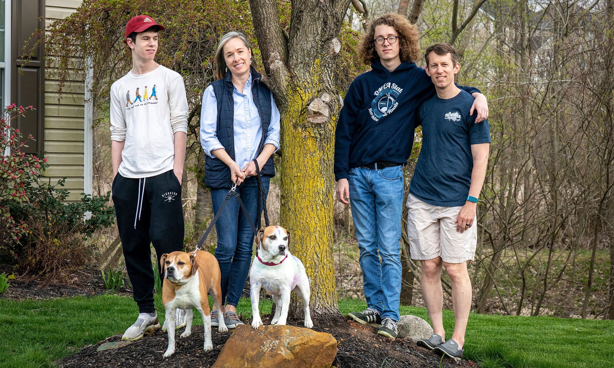 Four people stand outdoors in front of a tree, with two dogs sitting on a rock at their feet. The group appears relaxed and is dressed casually, with greenery and a house visible in the background.
