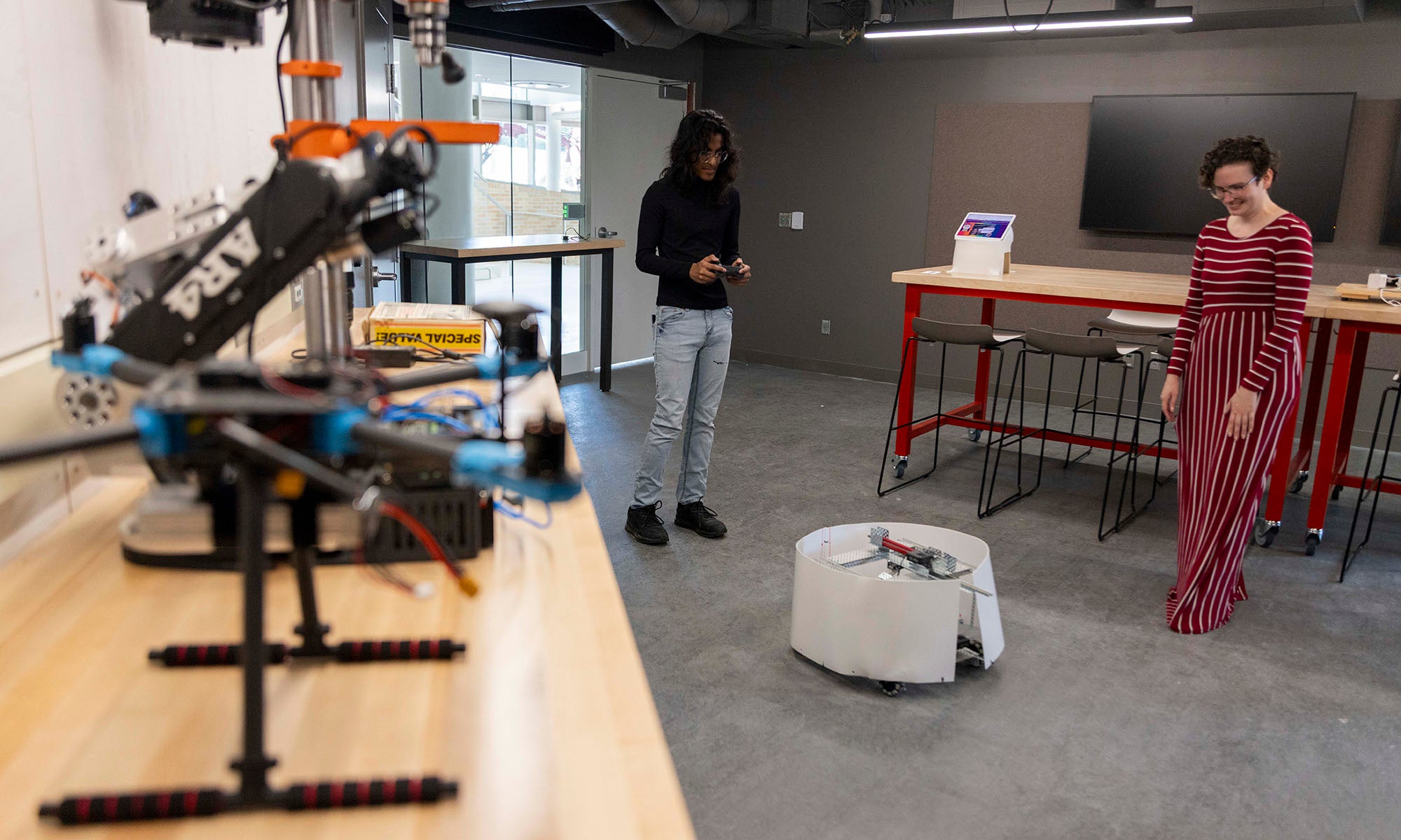 Two people observe a small wheeled robot in a workspace with tools, a workbench, and electronic equipment visible in the background.