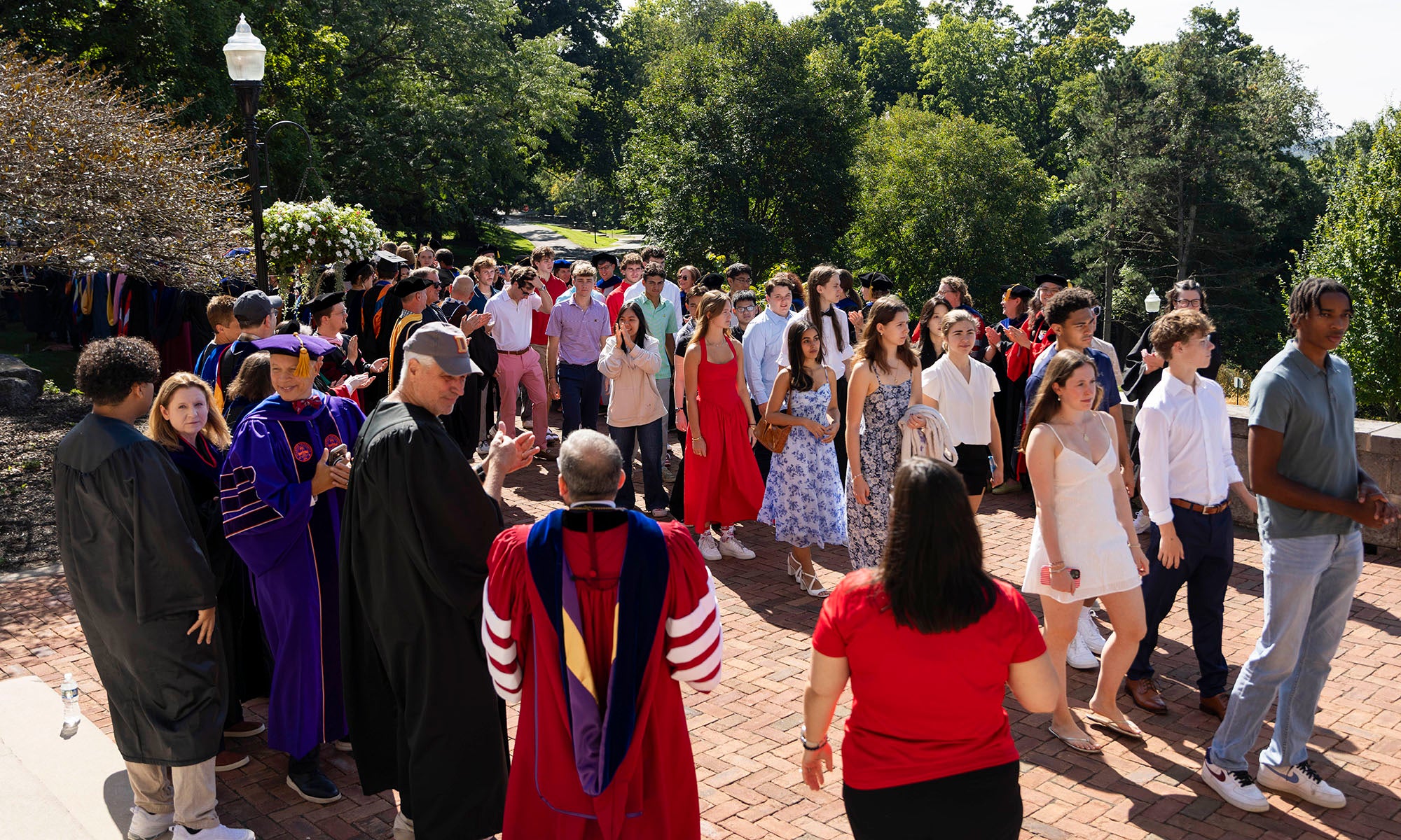 A group of students in semi-formal attire walk outdoors, while academic staff in robes stand nearby, during a daytime ceremony or event.