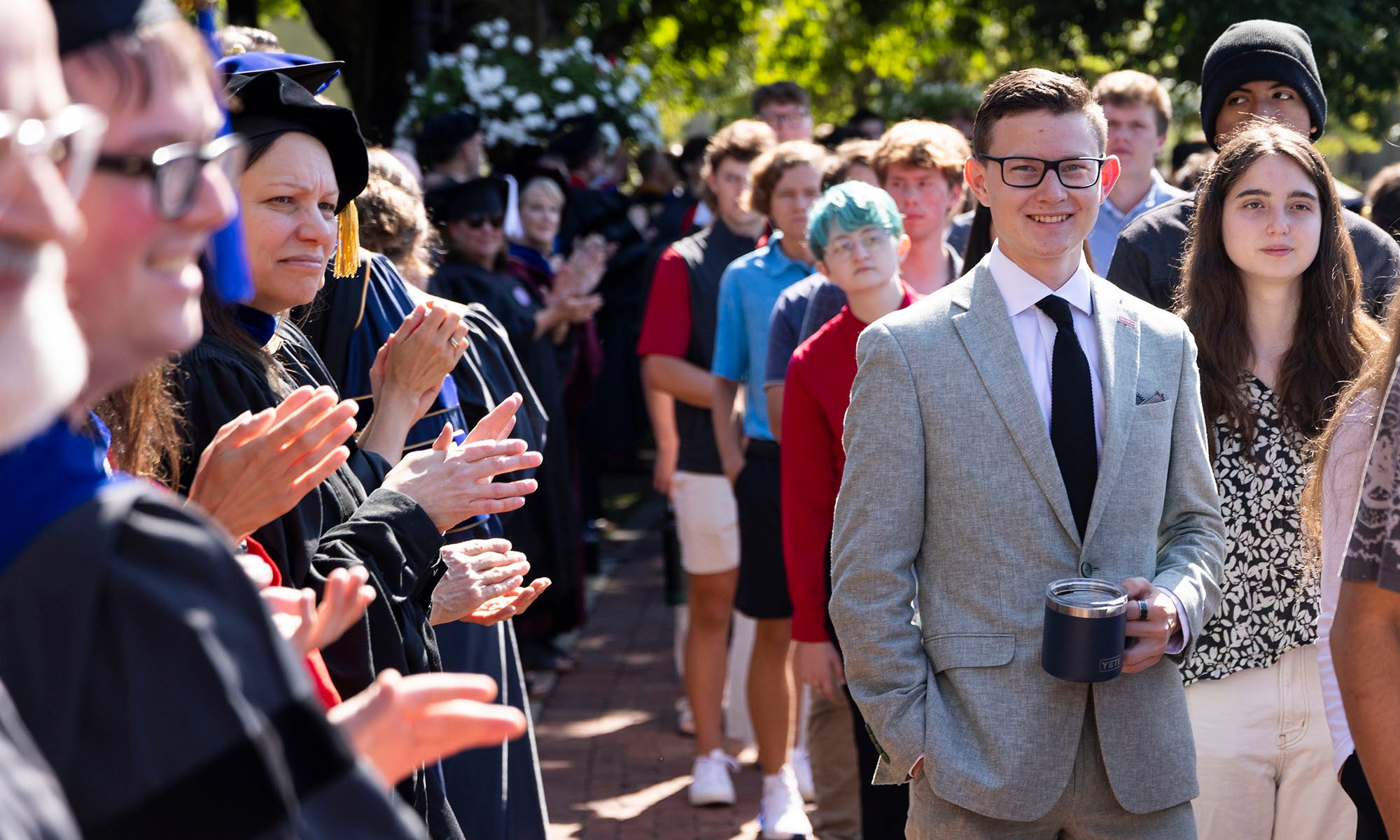 A group of people, some in graduation gowns, claps as others, including a man in a gray suit and a woman in patterned dress, walk past outdoors.