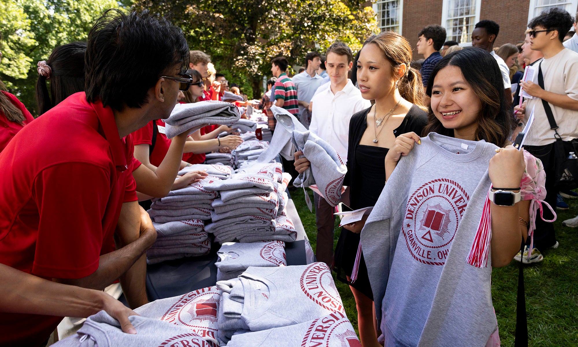 Students receive and hold up gray sweatshirts with a red university logo at an outdoor distribution table, while staff hand out more shirts.