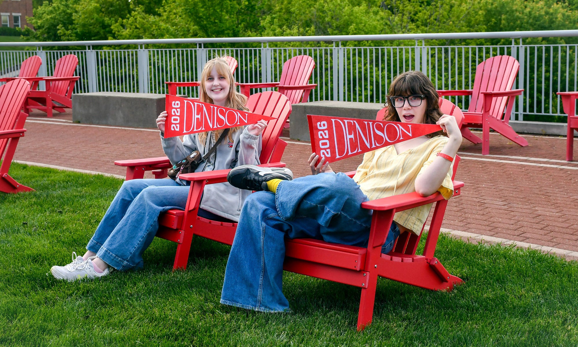 Two young women sit in red Adirondack chairs on a green lawn.