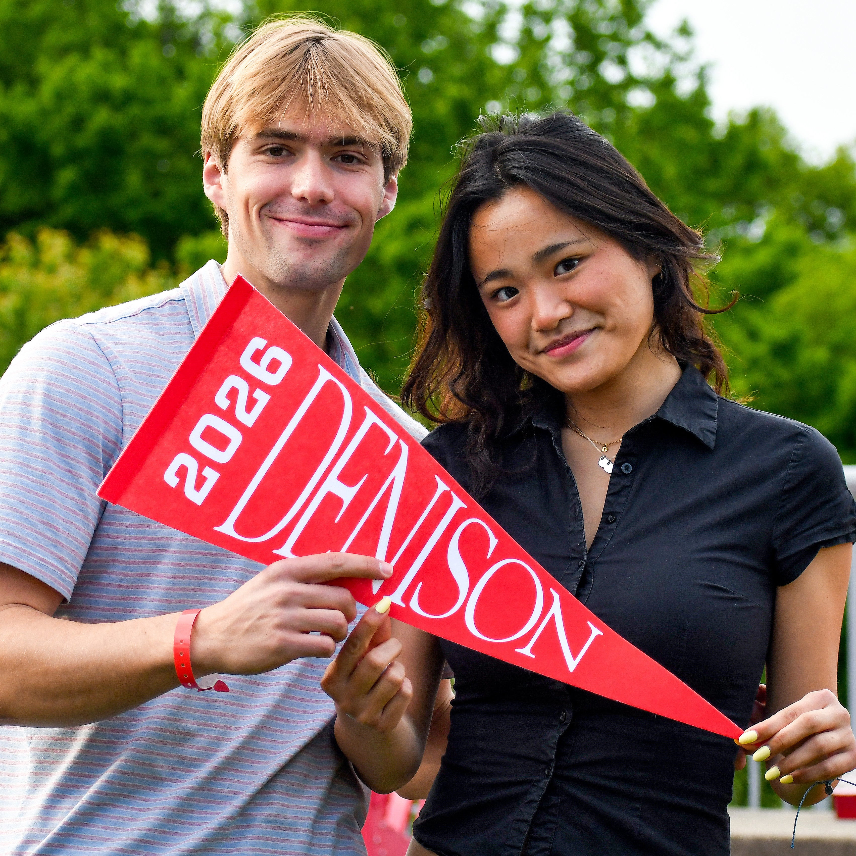 Two people stand outdoors holding a red pennant that says Denison 2026. Both are smiling and posing for the photo, with green trees in the background.