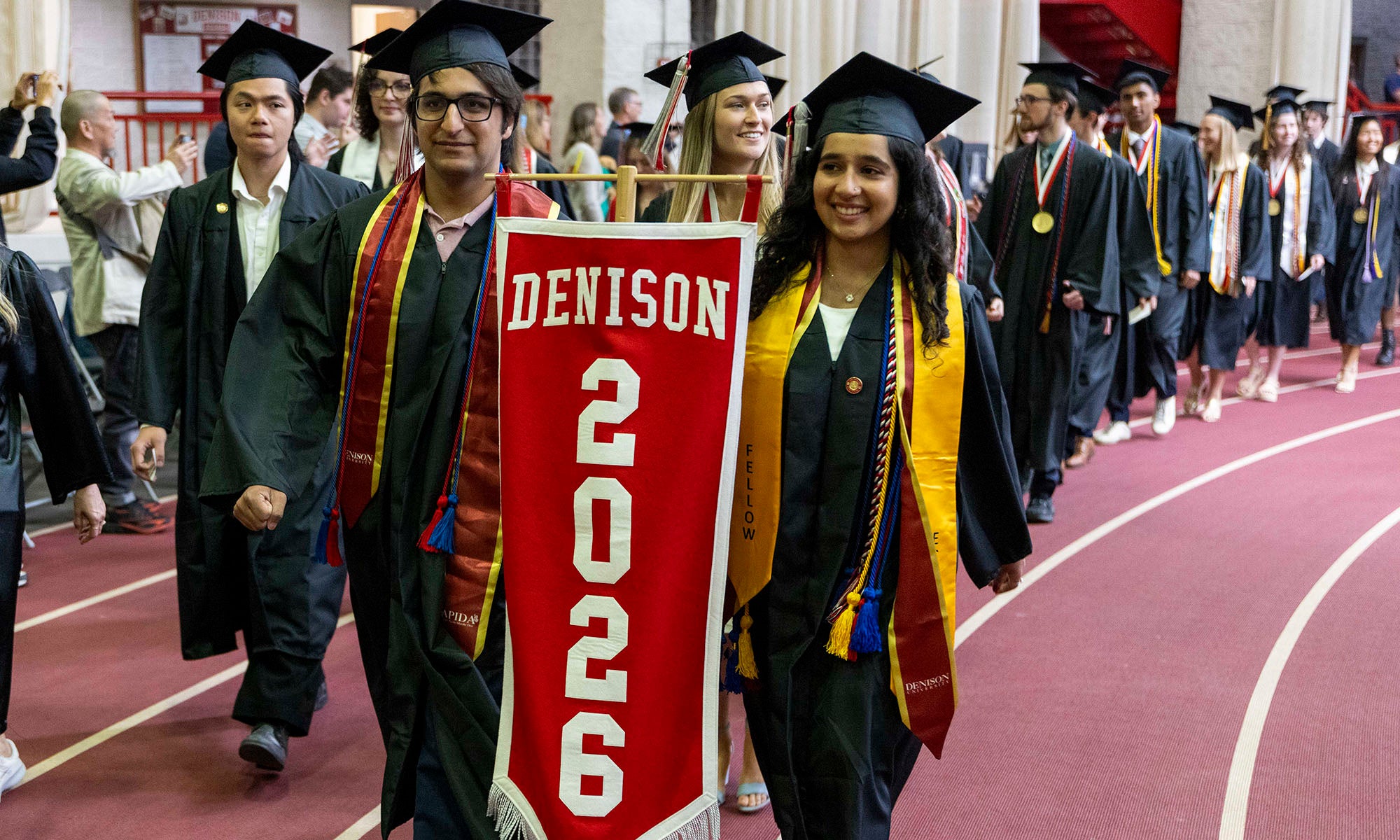 Two graduates carry a banner that reads "Denison 2026" as they lead a processional of seniors in graduation caps and gowns into a fieldhouse for a Commencement ceremony.