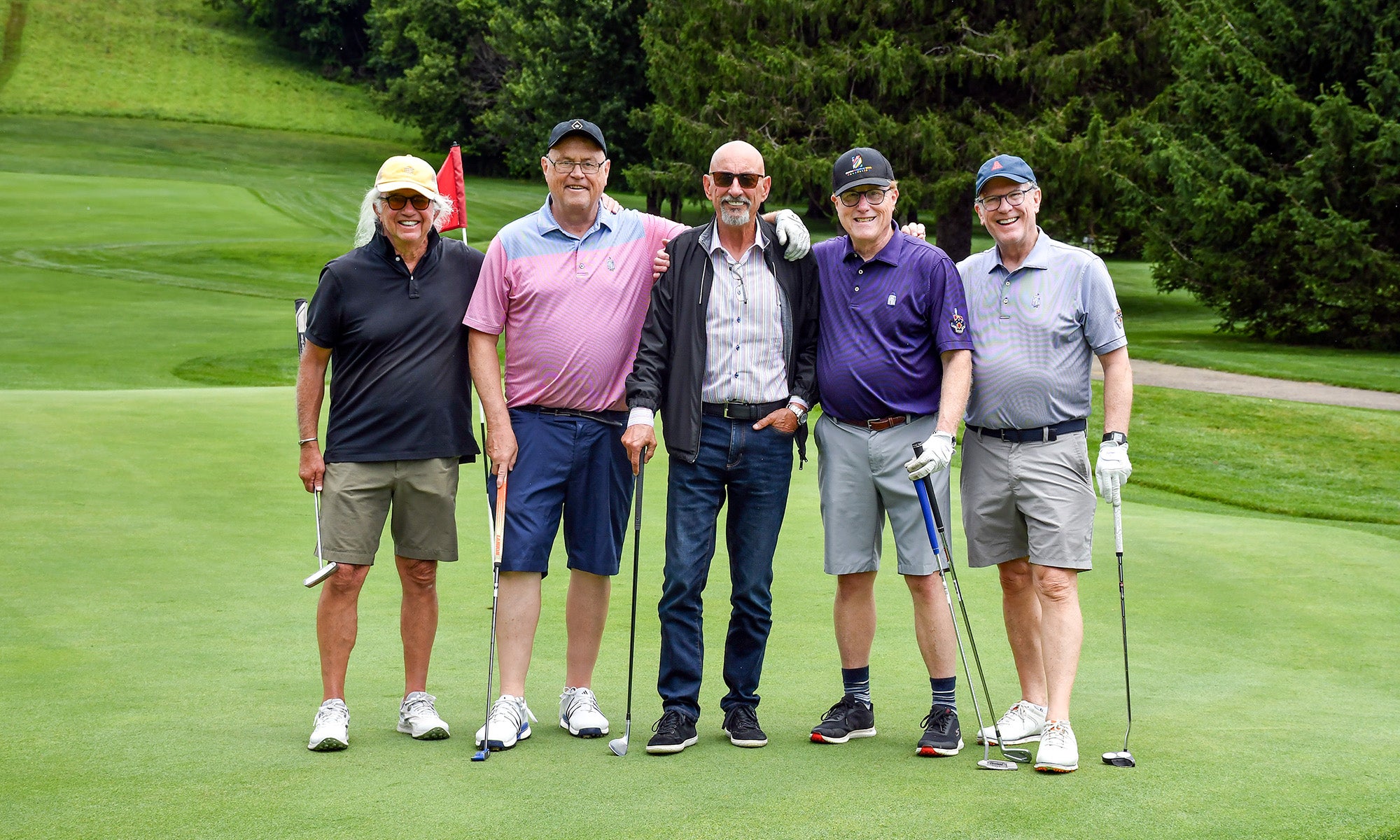 Five men stand together on a golf course, posing for a group photo with golf clubs and a flag visible in the background.