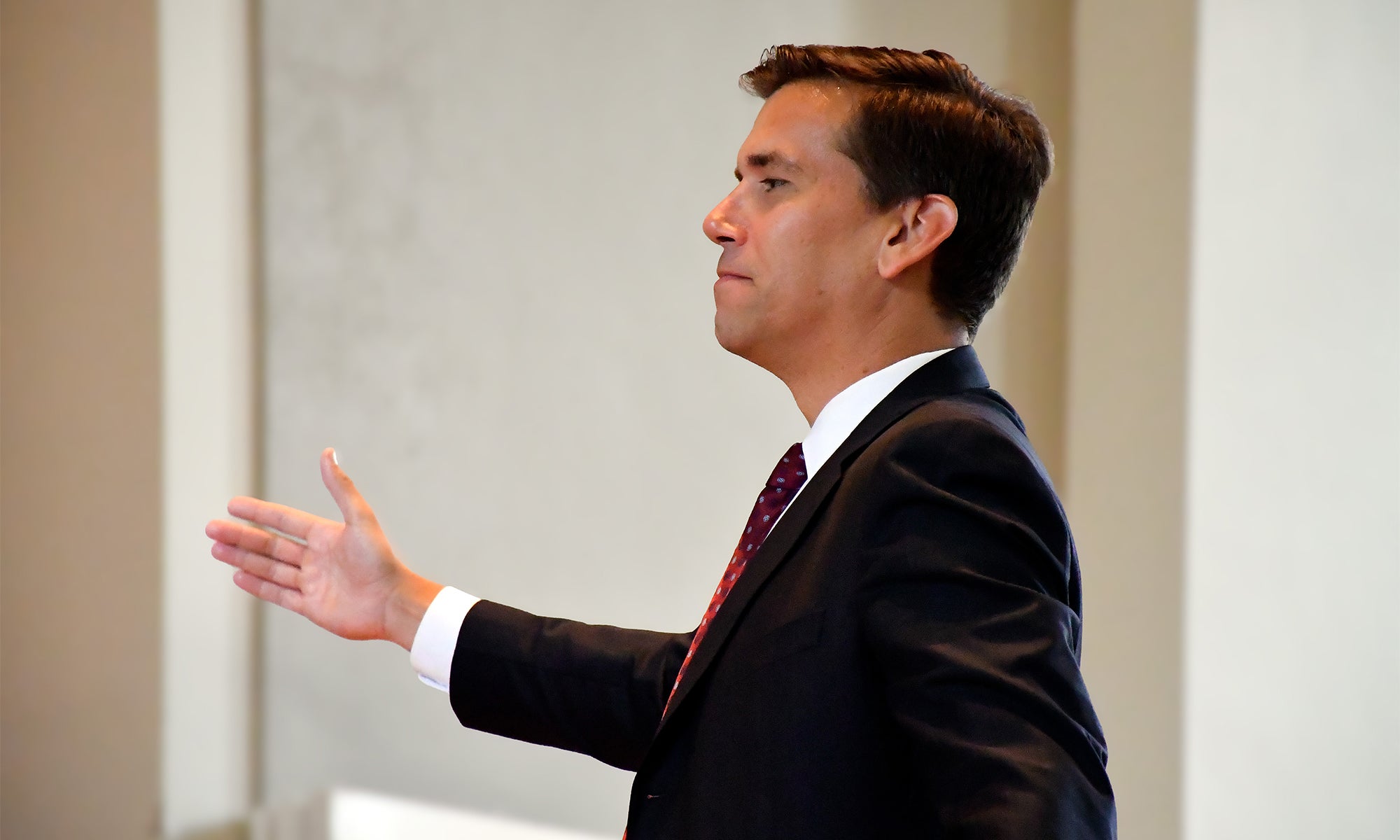 A man in a dark suit and red tie stands indoors, extending his right hand as if conducting a chorus. He is facing left in a well-lit room.
