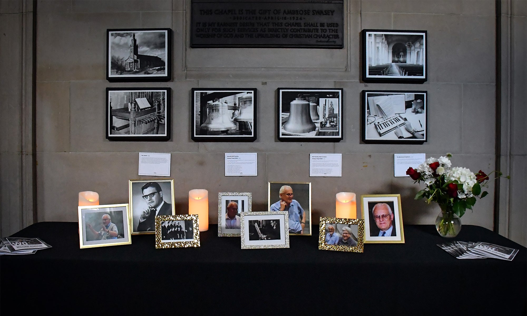 A memorial display with framed photos of various people, lit candles, and a bouquet of flowers on a black table. Black-and-white photographs and written notes are mounted on the wall behind the table.