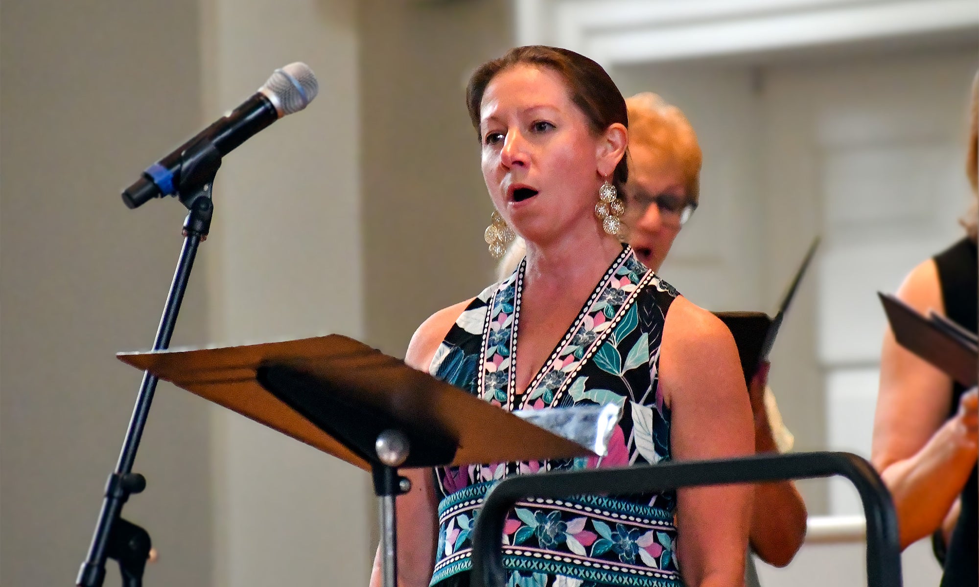 A woman stands singing into a microphone, holding sheet music on a stand, with two other women—members of the Denison Singers—singing and holding music in the background at a performance honoring Distinguished Professor Emeritus William Osborne.