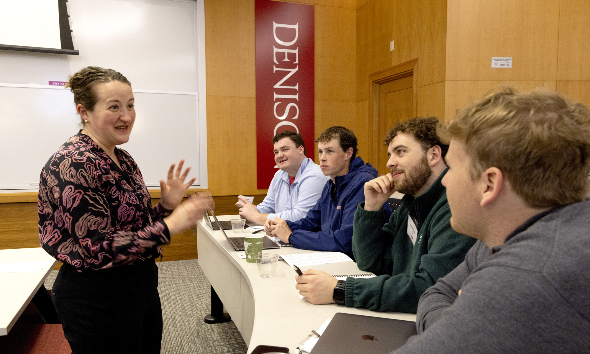 A woman speaks to four seated students in a classroom with a Denison banner on the wall; laptops and notebooks are on the table.