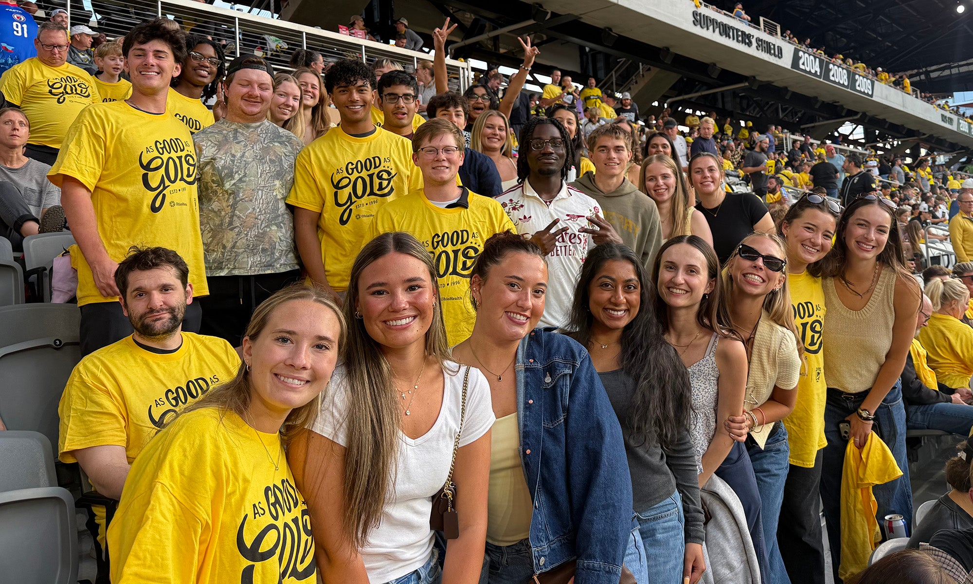 A group of people, many wearing yellow shirts, sit and stand together in stadium seats, smiling at the camera.