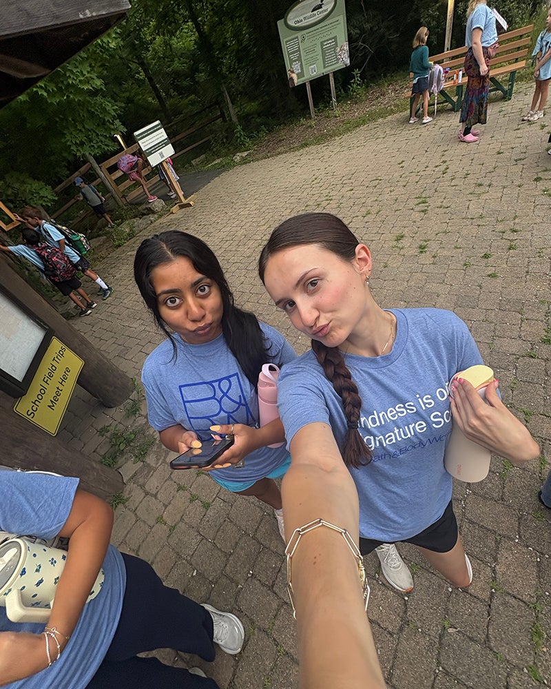 Two young women stand outdoors on a paved path, posing for a selfie. One holds a phone and water bottle, the other holds a snack. Both wear blue shirts. Other people are in the background.