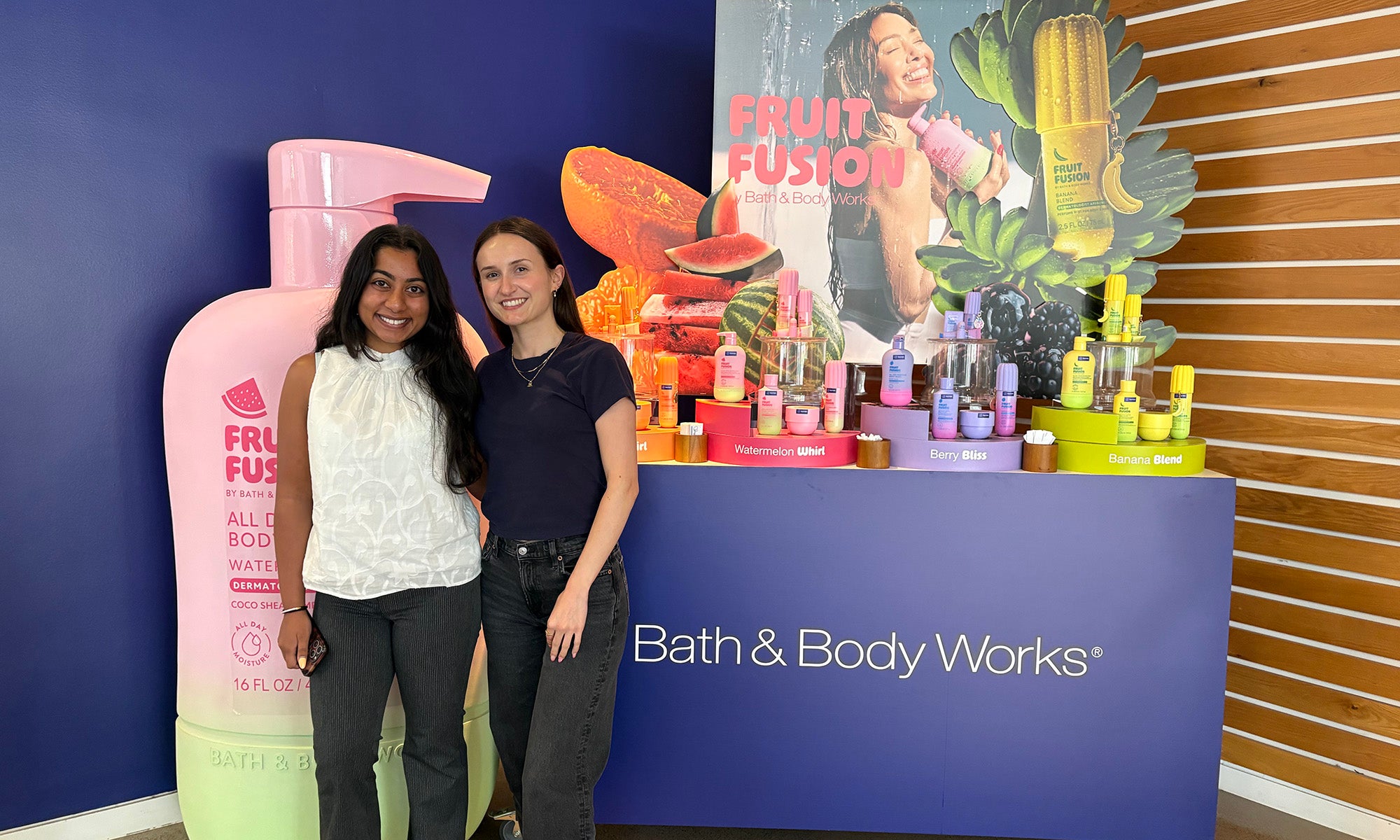 Two women stand smiling next to a Bath & Body Works display featuring colorful Fruit Fusion products and large promotional cutouts.