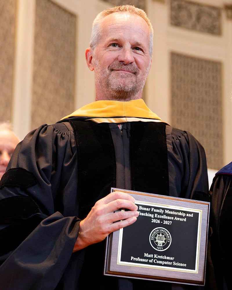 A man in doctoral academic regalia stands on a stage holding an academic award.