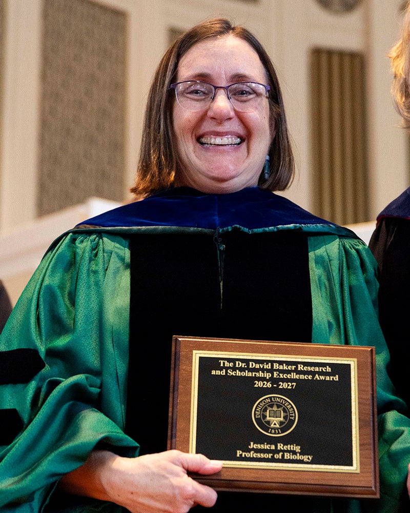 A woman in green and black academic regalia stands on a stage holding an academic award.