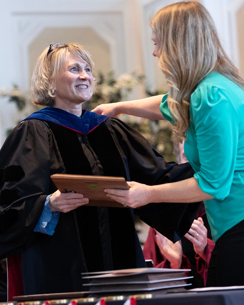 A woman in academic regalia holds a plaque as a woman in a green blouse presents it to her during a ceremony. They are standing in front of a blurred audience and floral arrangements.