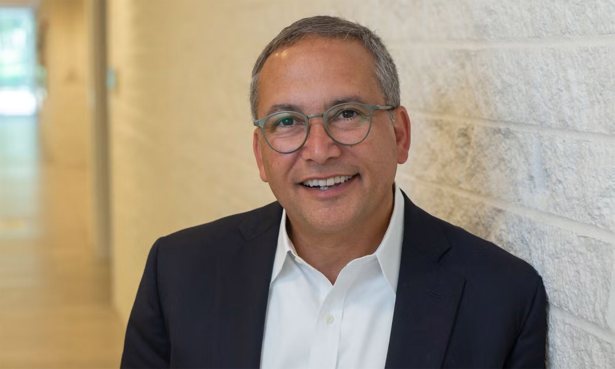 A man with short gray hair and glasses, wearing a white shirt and dark blazer, stands smiling against a light-colored brick wall in a hallway.