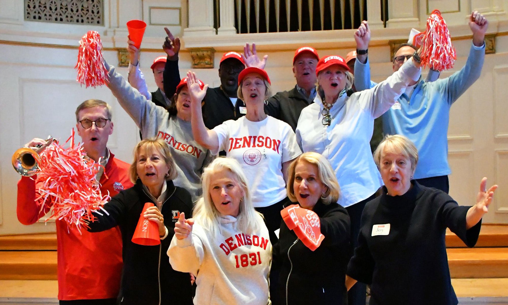 A group of adults wearing red and white clothing and hats stand indoors, holding pom-poms and red cups, cheering with energetic expressions. They are in front of wooden steps in a large hall.