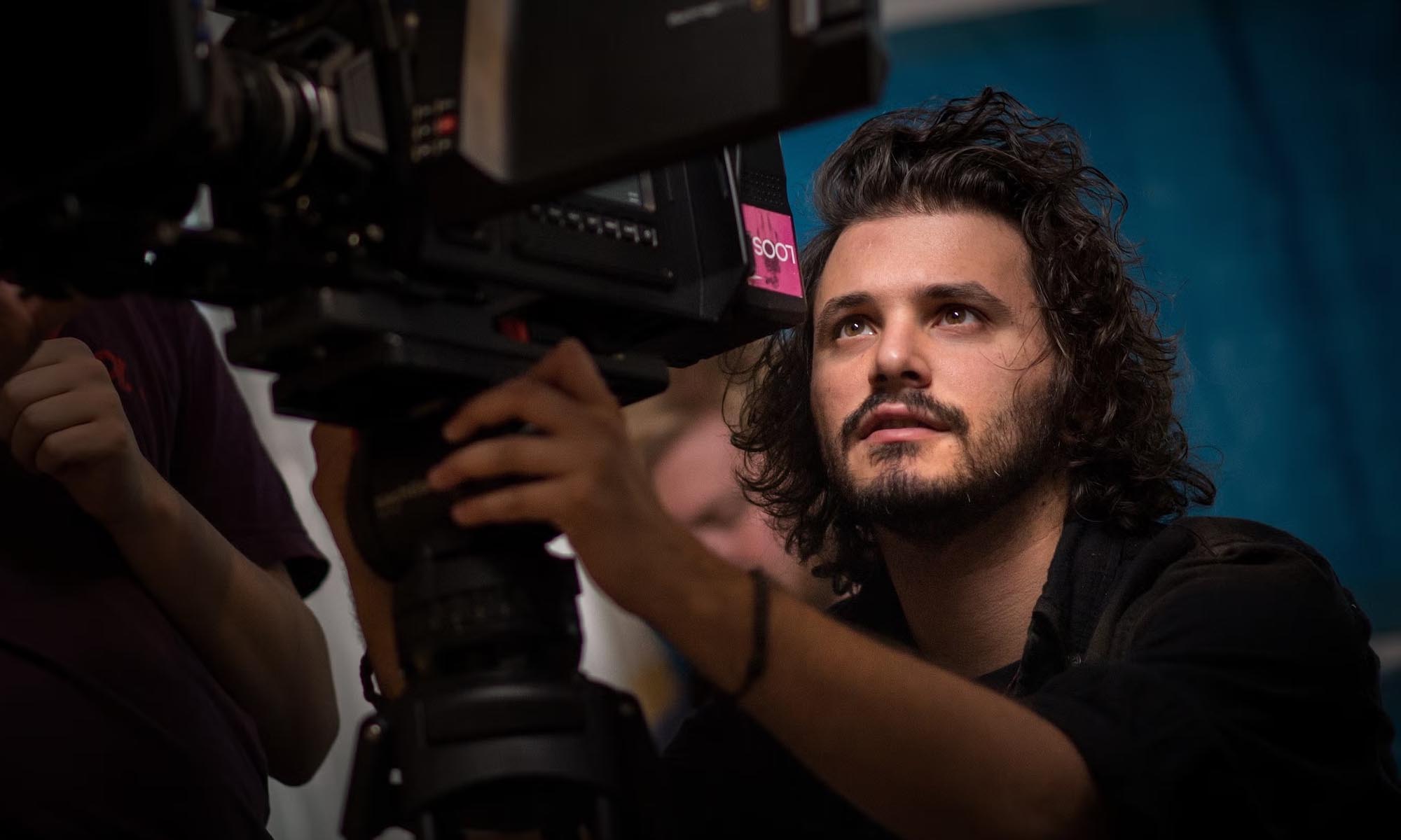 A man with curly hair operates a professional video camera, focusing intently, with a blurred person and blue background behind him.