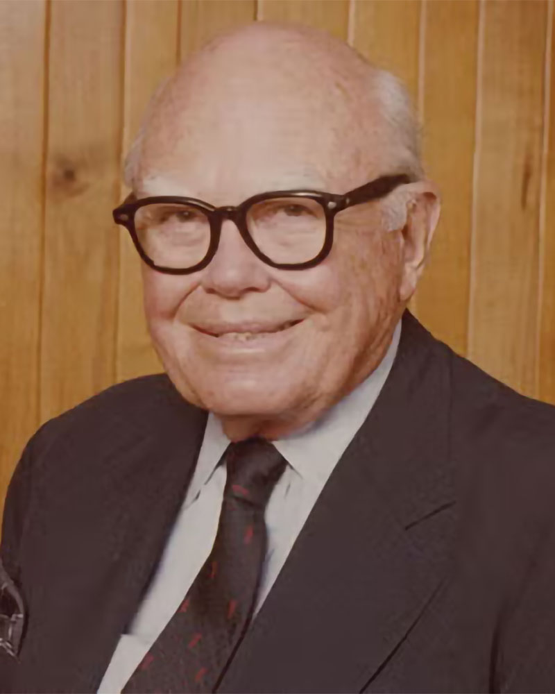 An older man with glasses, a suit, and a tie smiles in front of a wooden-paneled wall.