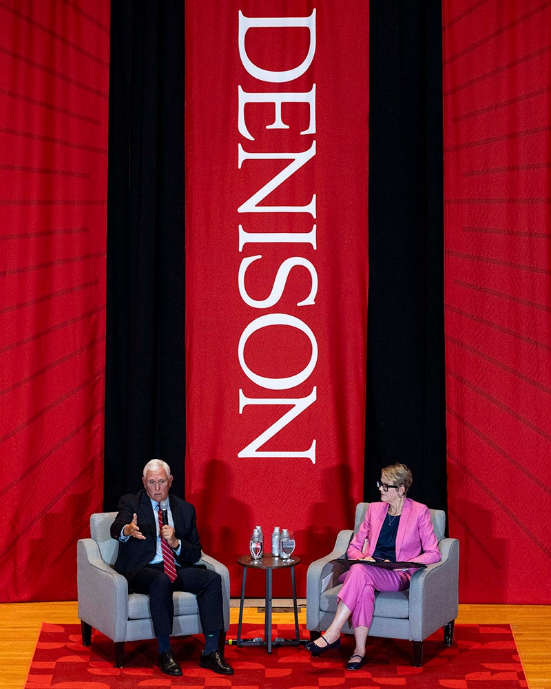 A man and woman sit in modern gray armchairs on a stage, flanked by tall red drapes and a Denison University banner.