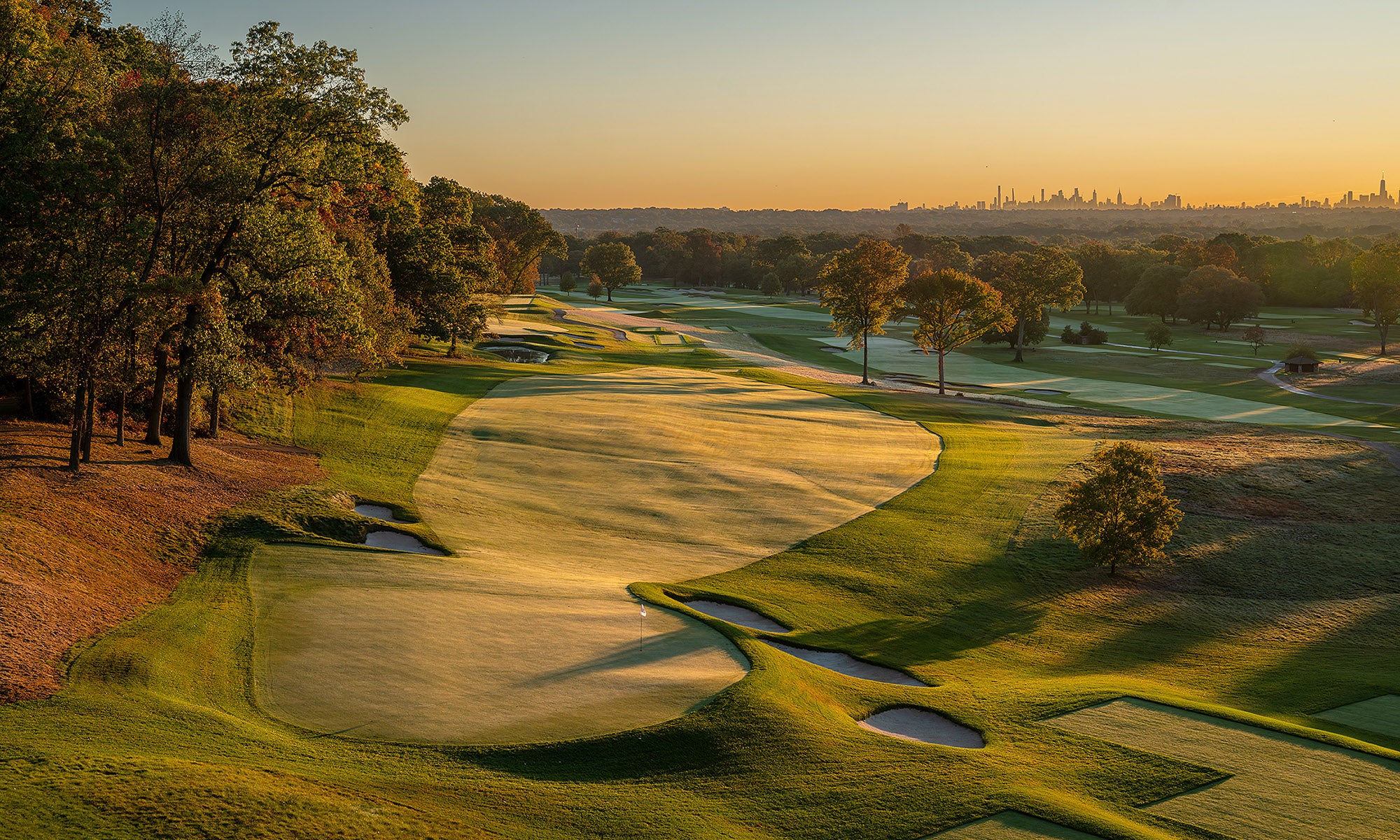 A golf course with sand bunkers and wide fairways is shown at sunrise, with trees lining the edges and a city skyline faintly visible in the background.