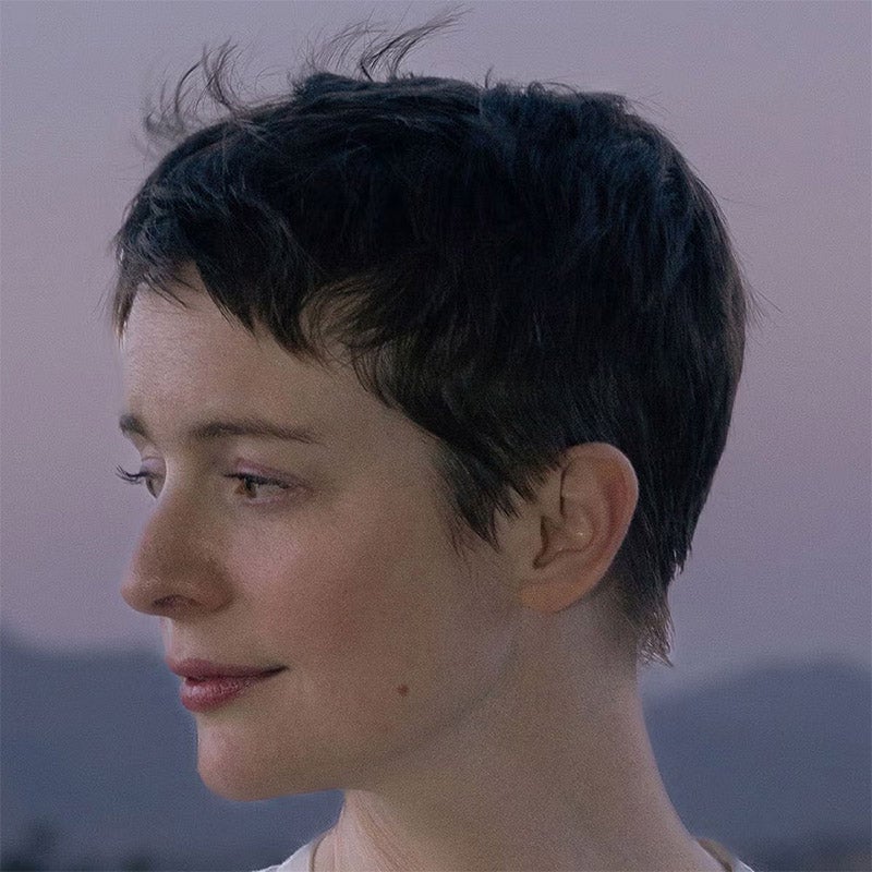 Person with short dark hair gazes to the left against a soft, blurred outdoor backdrop with pale sky.