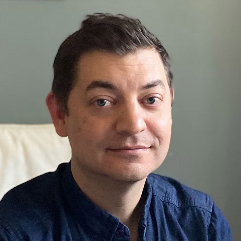 A man with short dark hair wearing a blue button-up shirt sits in front of a light gray wall, looking at the camera with a neutral expression.