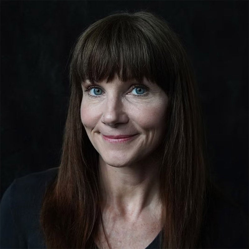 Woman with long brown hair and bangs, wearing a dark top, smiling slightly, posed against a dark background.