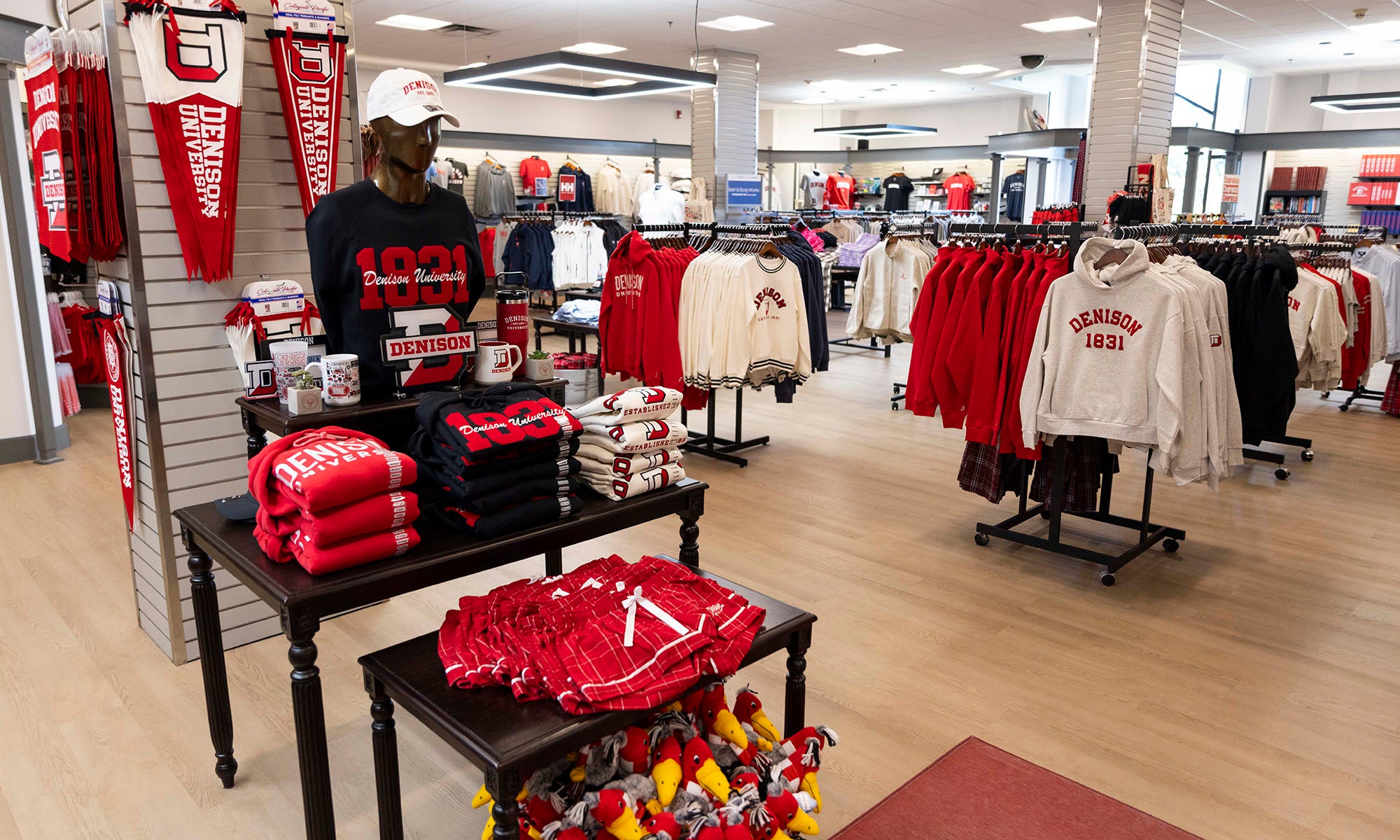 Campus bookstore interior with racks and tables displaying red and white university apparel, sweatshirts, hats, mugs, and other school merchandise. Bright, organized retail setting.
