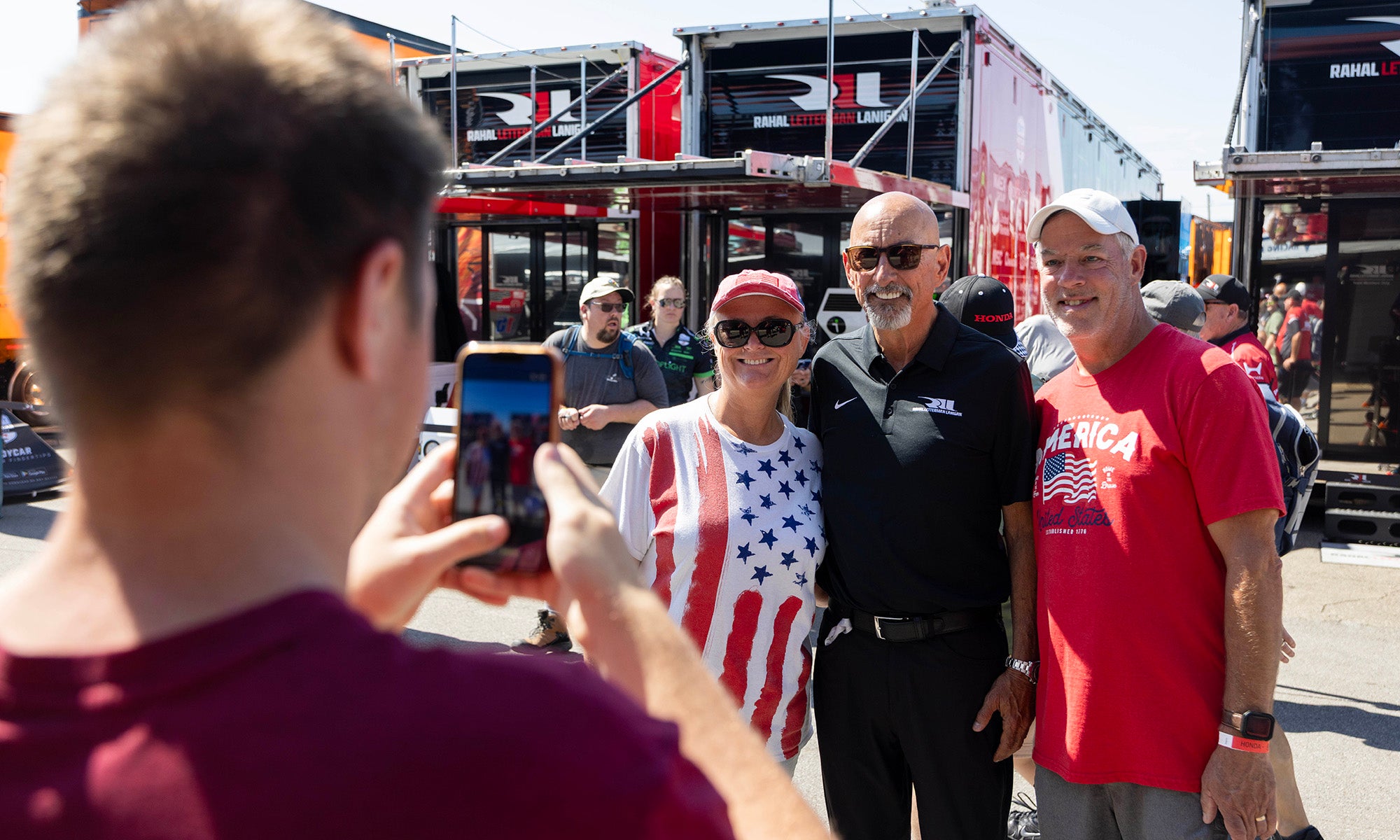 A person takes a photo of three adults posing together outdoors in front of large red trailers on a sunny day.
