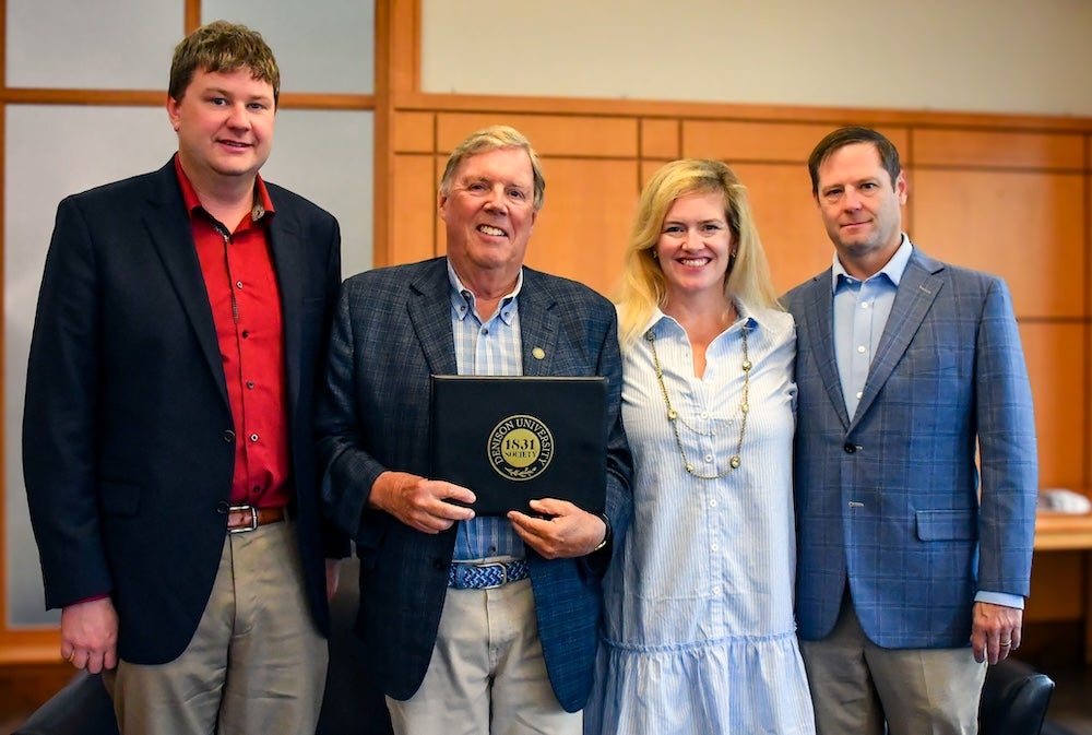 Four adults stand indoors, posing and smiling for the camera. One man in the center holds a folder with a gold seal. They are dressed in business or business casual attire.