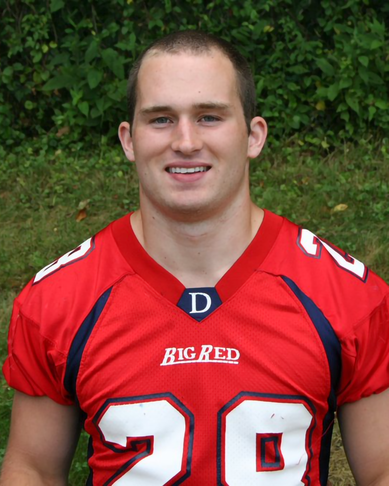 A young man wearing a red football jersey with the number 28 stands outdoors in front of greenery, smiling at the camera.