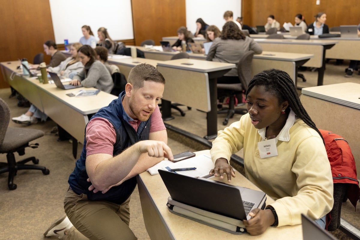 A man and a woman sit at a classroom desk, looking at a laptop together