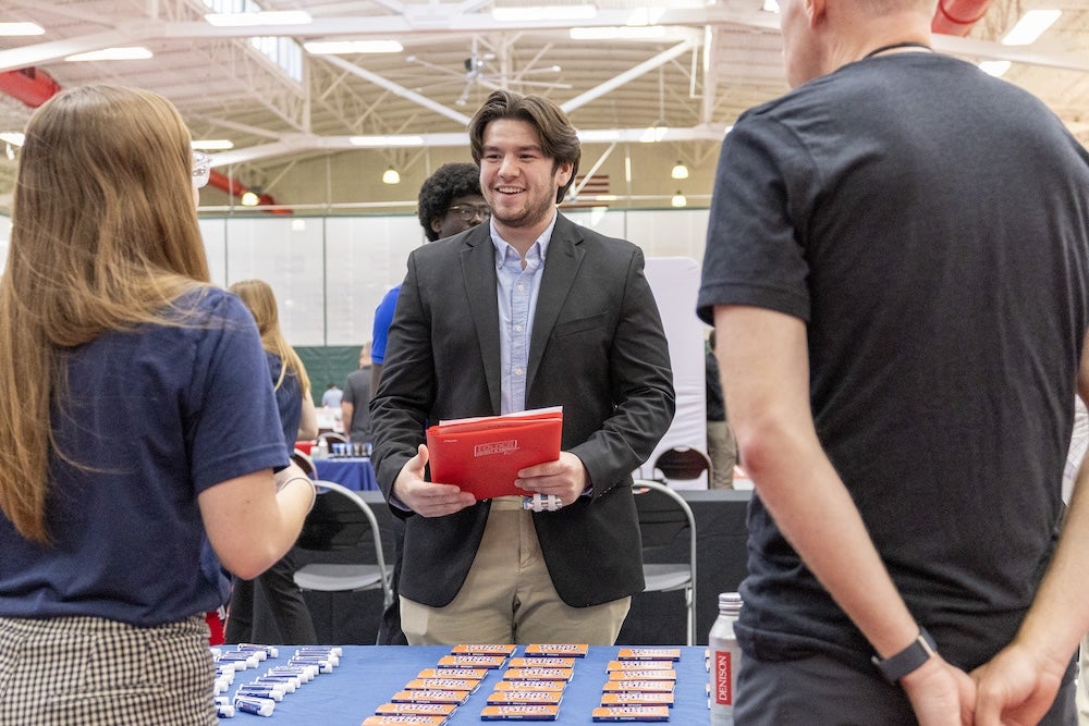 A man in a blazer holding a red folder talks to two people at a table during the career expo.
