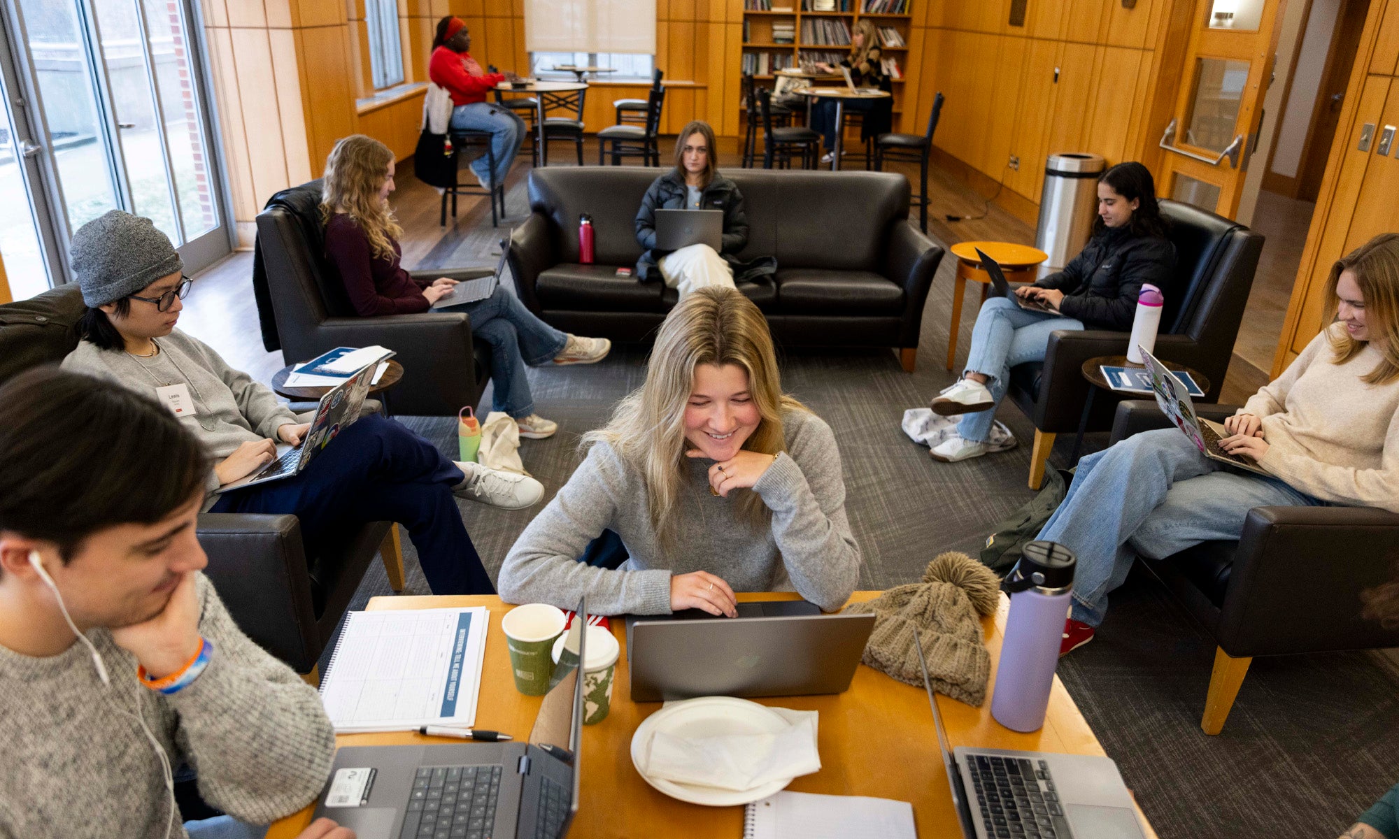 A modern wood- and glass-paneled meeting room filled with college students on laptops seated at tables, couches, and chairs.