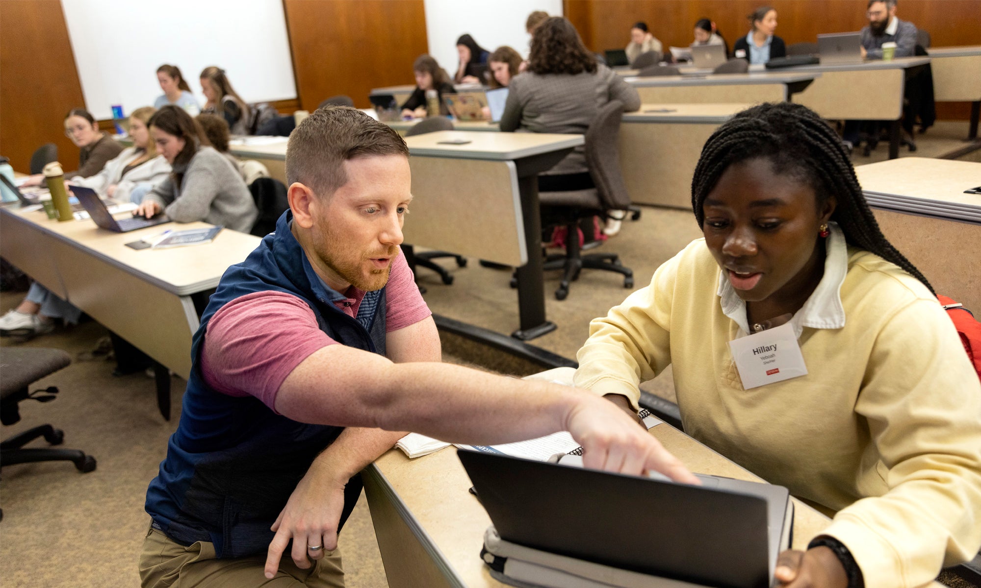 A staff member reviews a student's work on a laptop as other students and staff meet in groups in the background.