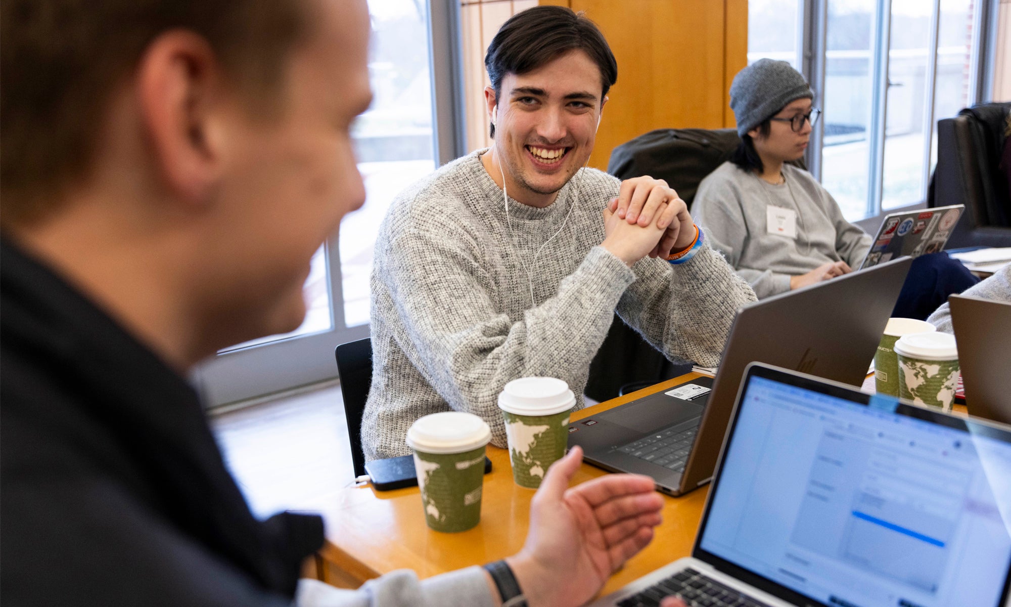 A student listening and smiling during a discussion with other students around a large conference table in front of a large bank of windows.